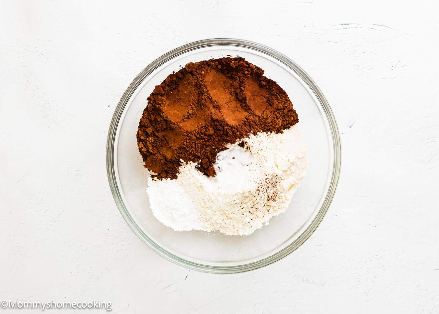 A glass bowl containing cocoa powder, flour, baking powder, baking soda, and salt on a white surface—perfect for making Ultimate Chocolate Cookies (Egg-Free).