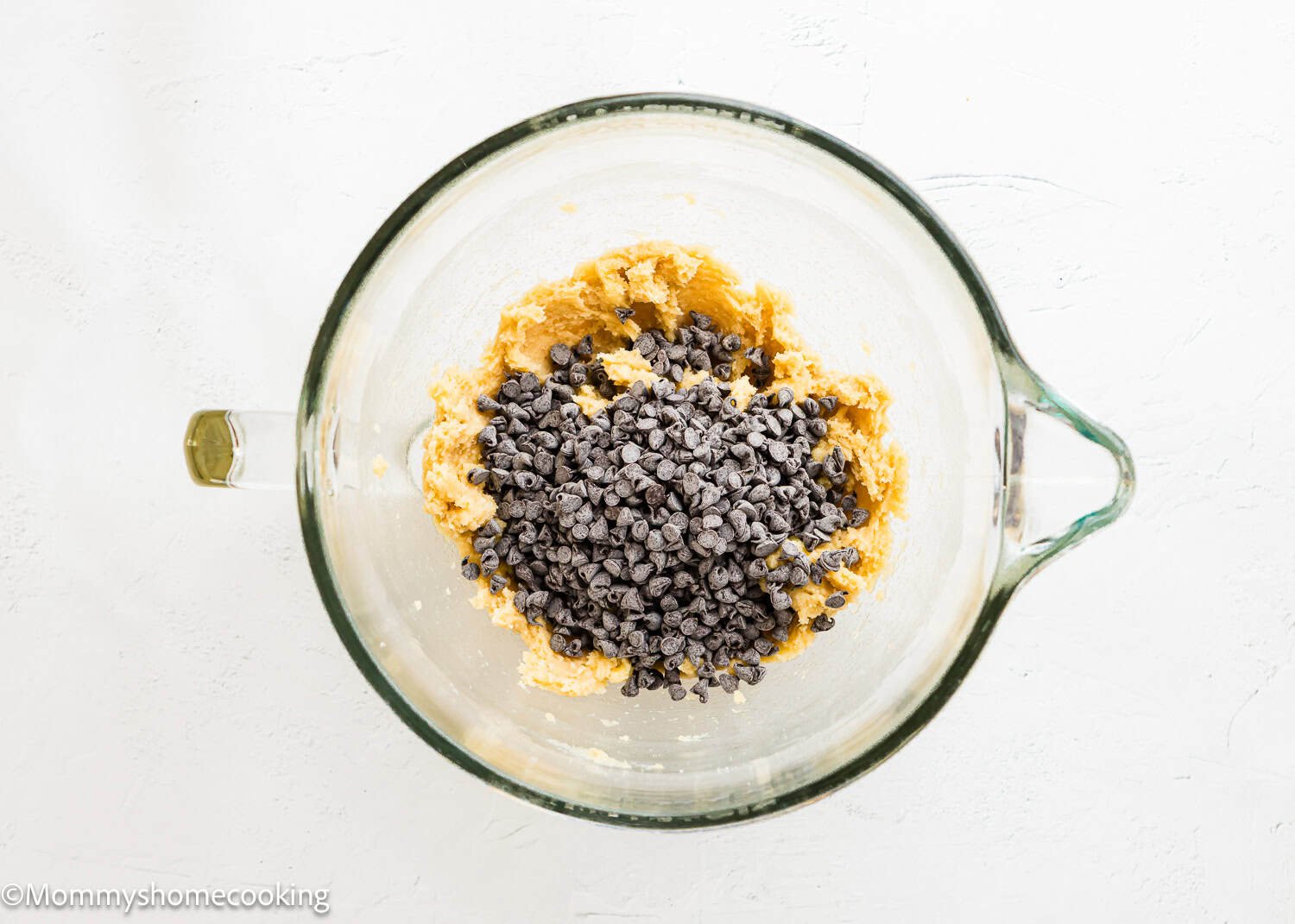 A glass mixing bowl containing Ultimate Chocolate Cookies (Egg-Free) dough with chocolate chips on top, placed on a white surface.