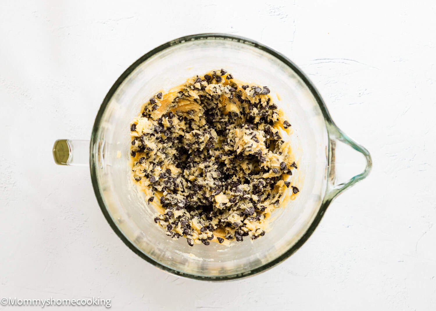A glass mixing bowl containing Ultimate Chocolate Cookies (Egg-Free) dough with chocolate chips sits on a white surface.