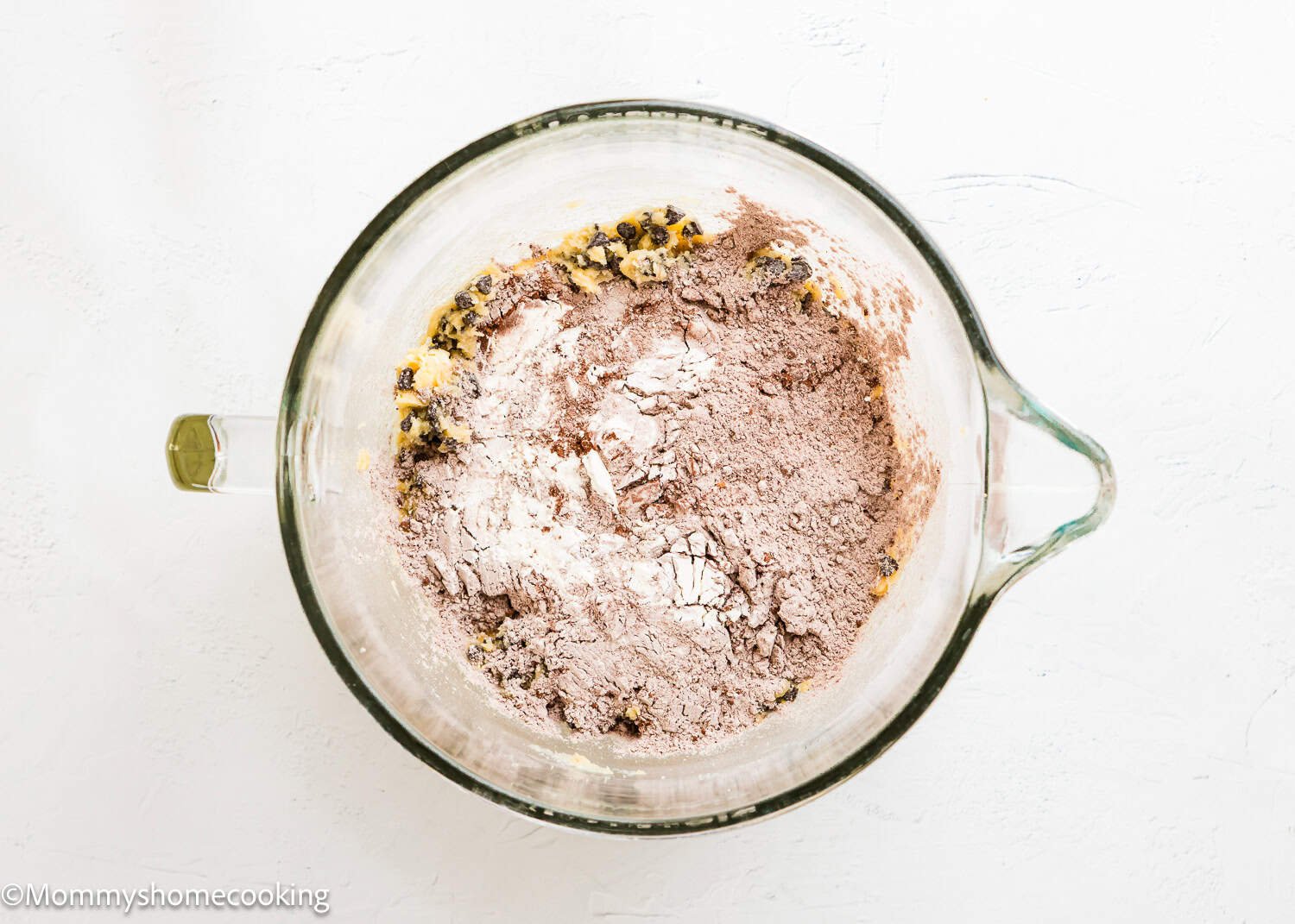 A glass bowl containing partially mixed Ultimate Chocolate Cookies (Egg-Free) dough with visible flour, cocoa powder, and chocolate chips on a white surface.