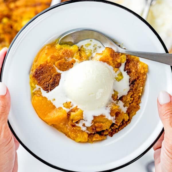 A close-up of Ultimate Easy Peach Dump Cake (5 Ingredients) on a plate, topped with a scoop of melting vanilla ice cream, held by two hands with a spoon on the side.