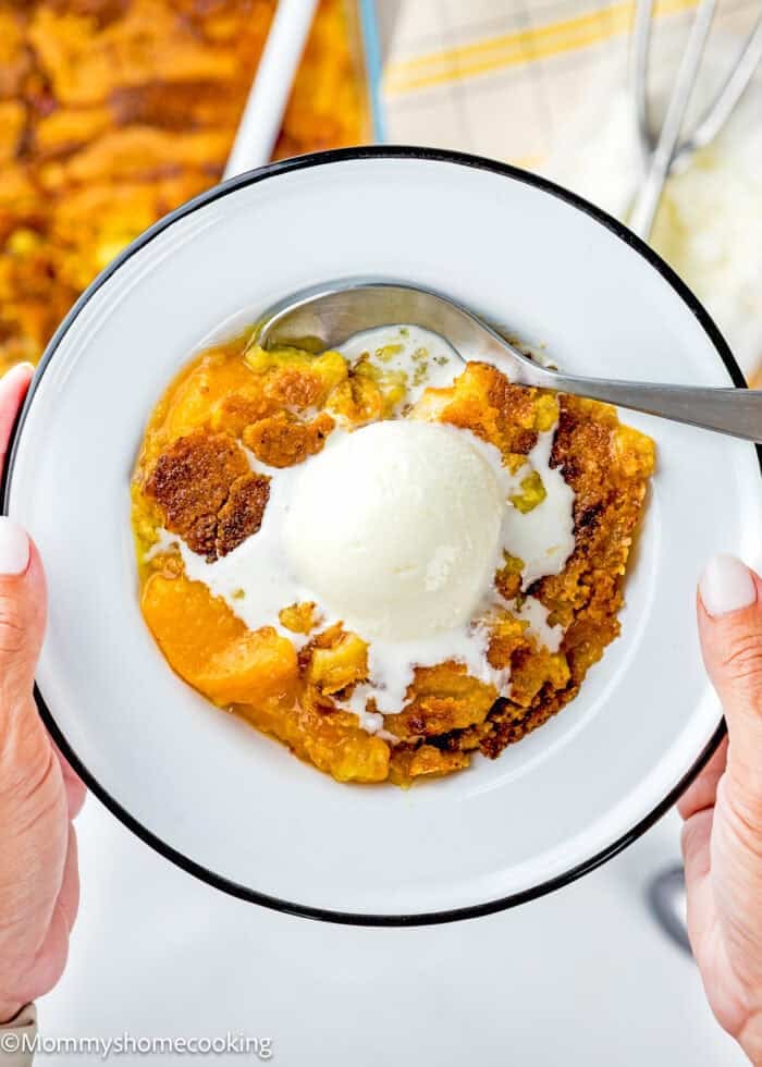 A close-up of Ultimate Easy Peach Dump Cake (5 Ingredients) on a plate, topped with a scoop of melting vanilla ice cream, held by two hands with a spoon on the side.