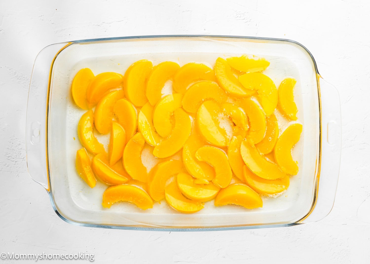 Glass baking dish with evenly spread peach slices on a white surface, ready for the Ultimate Easy Peach Dump Cake (5 Ingredients).