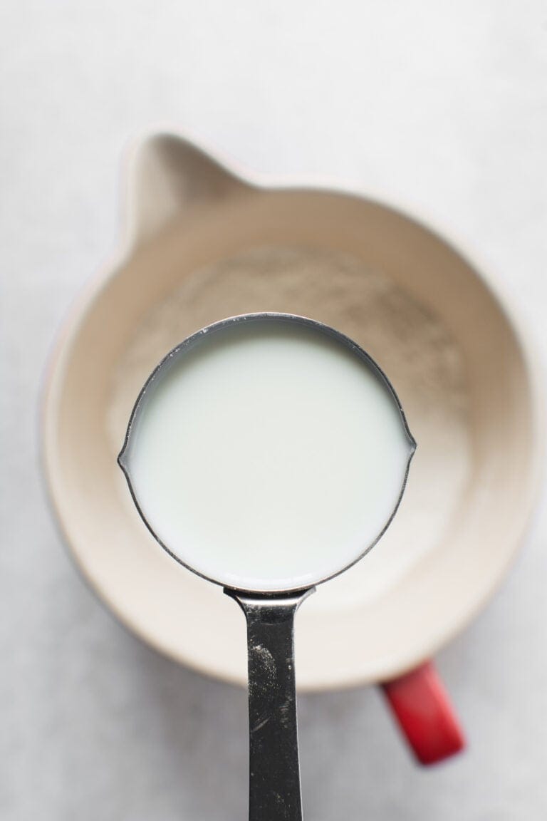 A metal measuring cup filled with milk is held above a mixing bowl of flour—perfect for trying Your Guide to Dairy-Free Swaps: How I Replace Dairy in My Family’s Egg-Free Recipes.