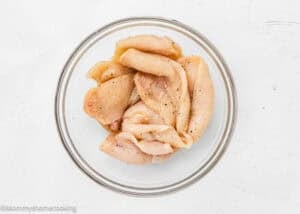 Raw, seasoned chicken breast strips ideal for Chicken Parmesan without Eggs are placed in a clear glass bowl on a white surface.