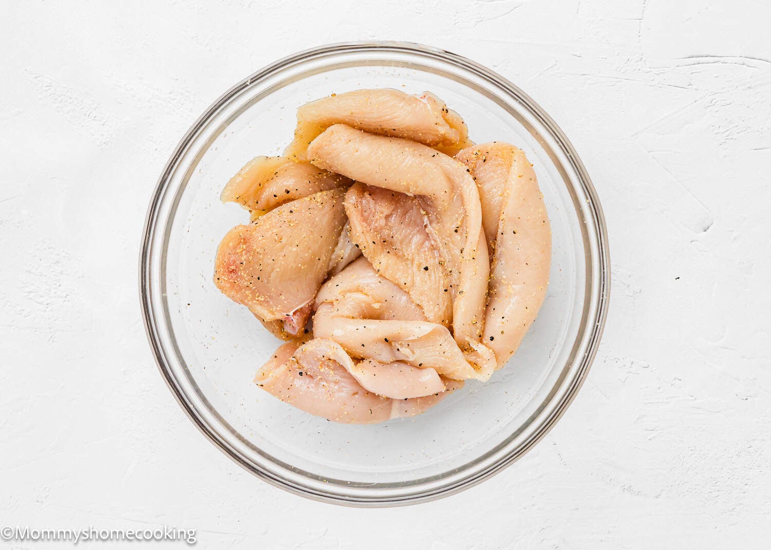 Raw, seasoned chicken breast strips ideal for Chicken Parmesan without Eggs are placed in a clear glass bowl on a white surface.
