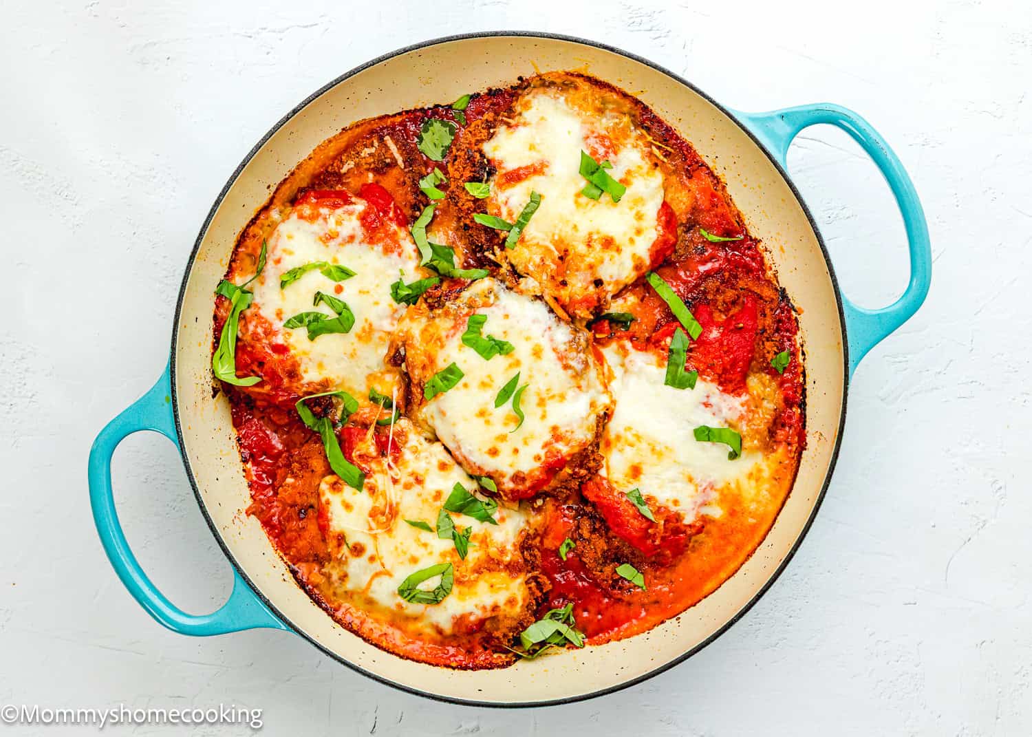 A blue skillet filled with baked Chicken Parmesan without Eggs, topped with melted cheese, tomato sauce, and fresh basil on a white surface.