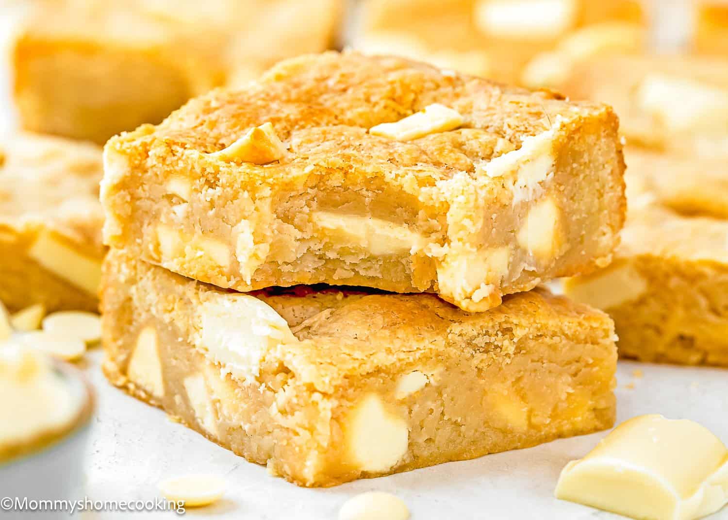 Two thick blondie bars without eggs with visible chunks of white chocolate are stacked on top of each other, surrounded by more blondies and pieces of white chocolate.