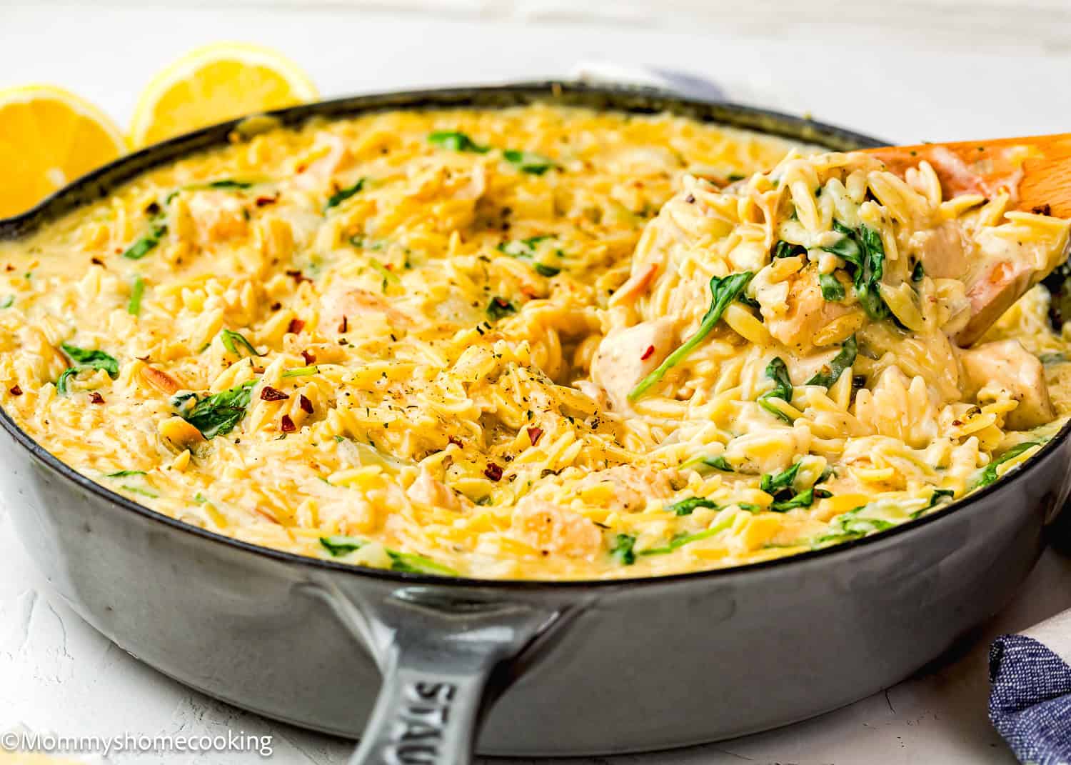 A skillet filled with creamy Easy Lemon Chicken Orzo (One Pot), mixed with spinach and tender chicken, as a wooden spoon lifts a serving. Lemon halves are visible in the background.