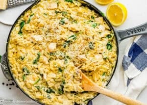 A skillet filled with Easy Lemon Chicken Orzo (One Pot), creamy orzo pasta, spinach, and chicken, topped with grated cheese, with a wooden spoon and lemon halves nearby.