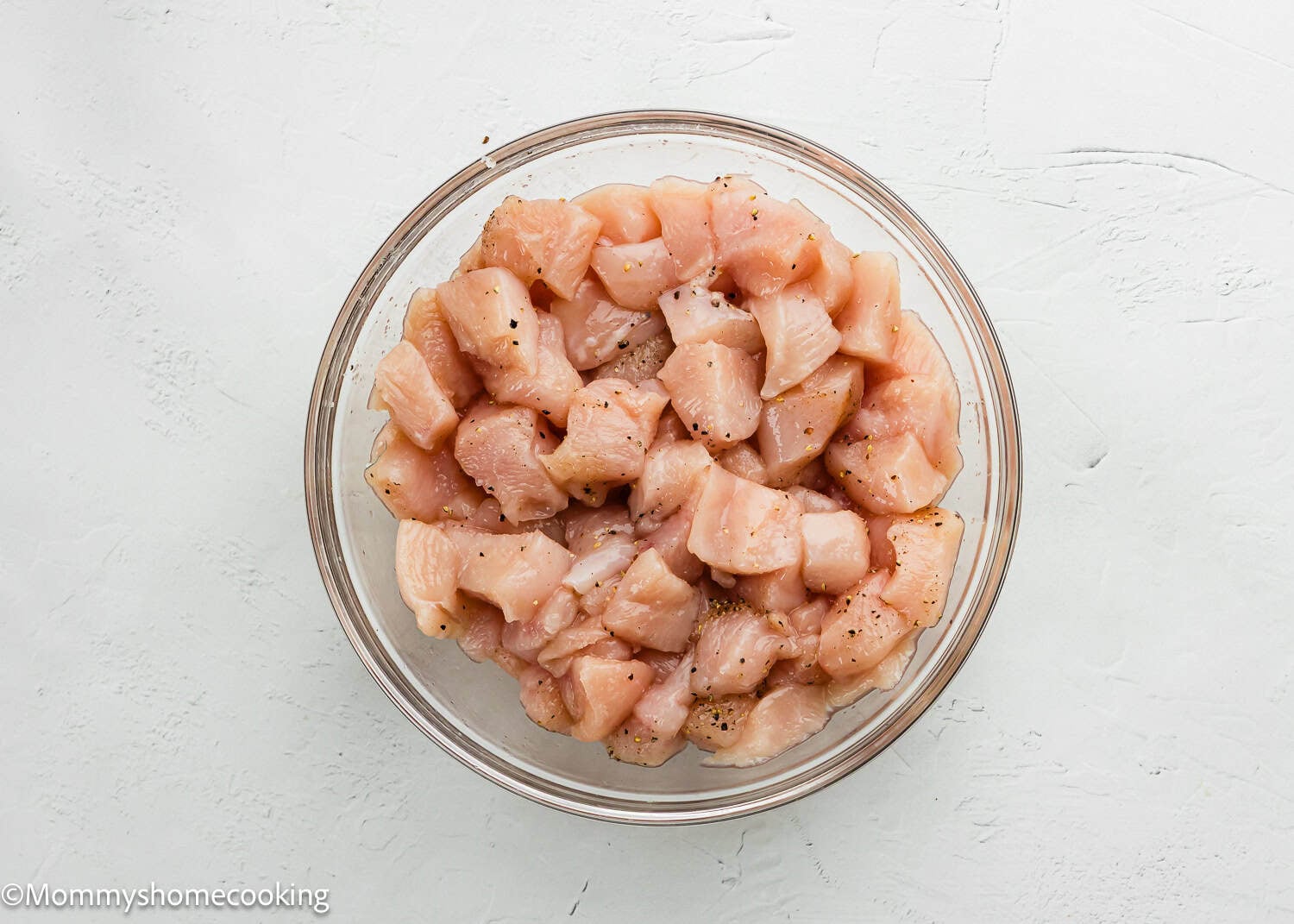 A glass bowl filled with raw, diced chicken breast pieces seasoned with pepper, perfect for preparing Easy Lemon Chicken Orzo (One Pot), set on a white textured surface.