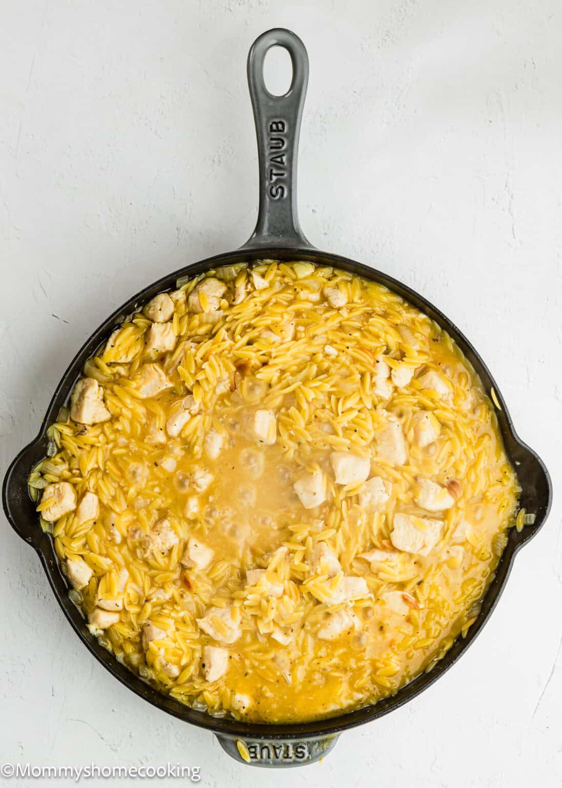 A cast iron skillet filled with Easy Lemon Chicken Orzo (One Pot) showcases cooked orzo pasta and chunks of chicken in a creamy sauce, viewed from above on a white background.