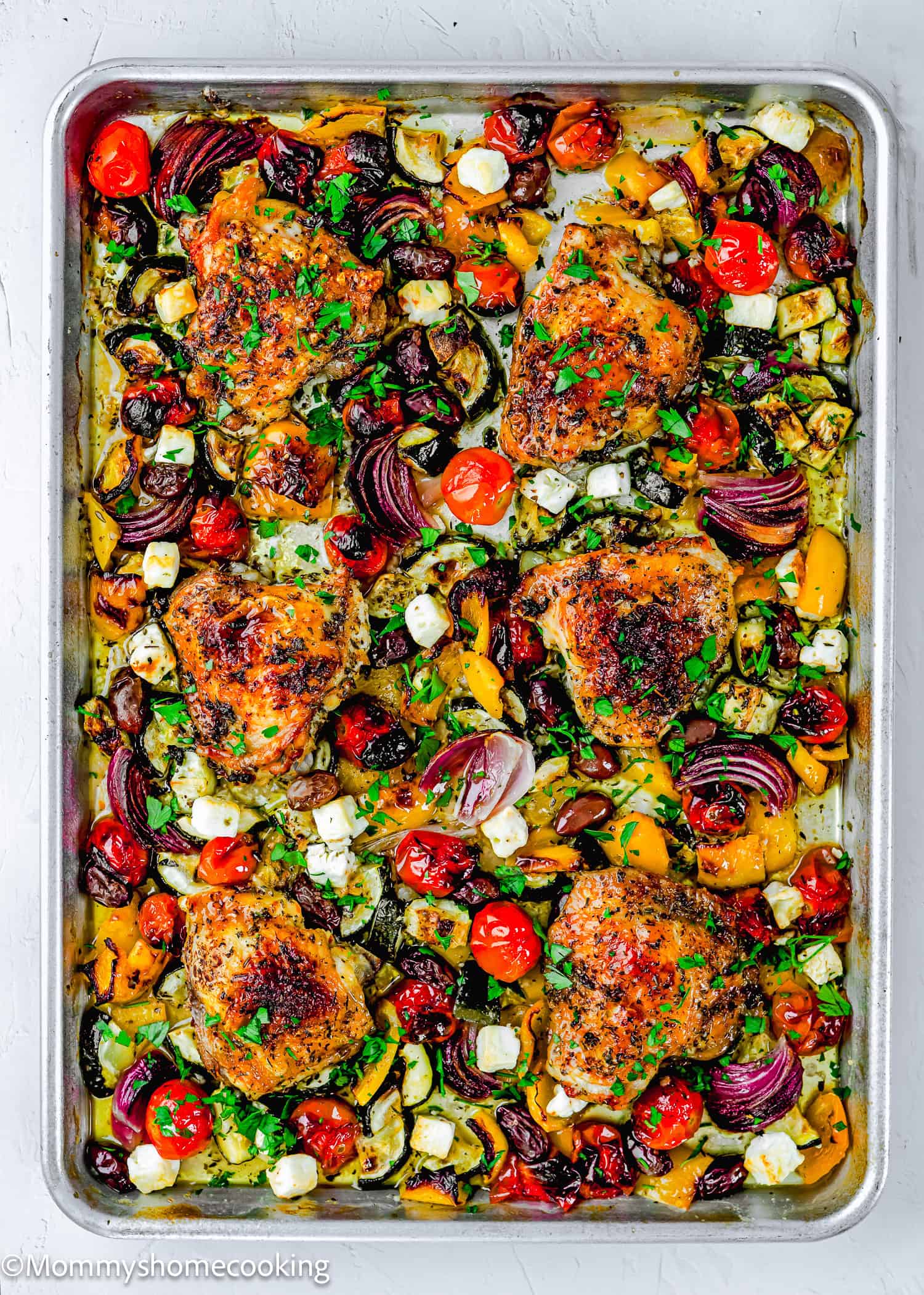 An Easy Mediterranean Sheet-Pan Chicken dinner featuring six seasoned chicken thighs surrounded by roasted cherry tomatoes, red onions, bell peppers, zucchini, and feta cheese, all garnished with chopped parsley.