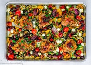 Easy Mediterranean Sheet-Pan Chicken with roasted chicken thighs, cherry tomatoes, red onions, zucchini, bell peppers, cubes of cheese, and fresh herbs on a white background.