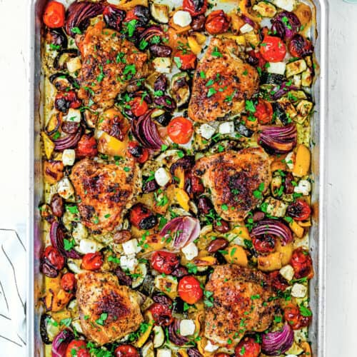 Easy Mediterranean Sheet-Pan Chicken: Roasted chicken thighs, red onions, zucchini, bell peppers, cherry tomatoes, and cubes of feta cheese garnished with fresh herbs—all on one pan for a delicious and effortless meal.
