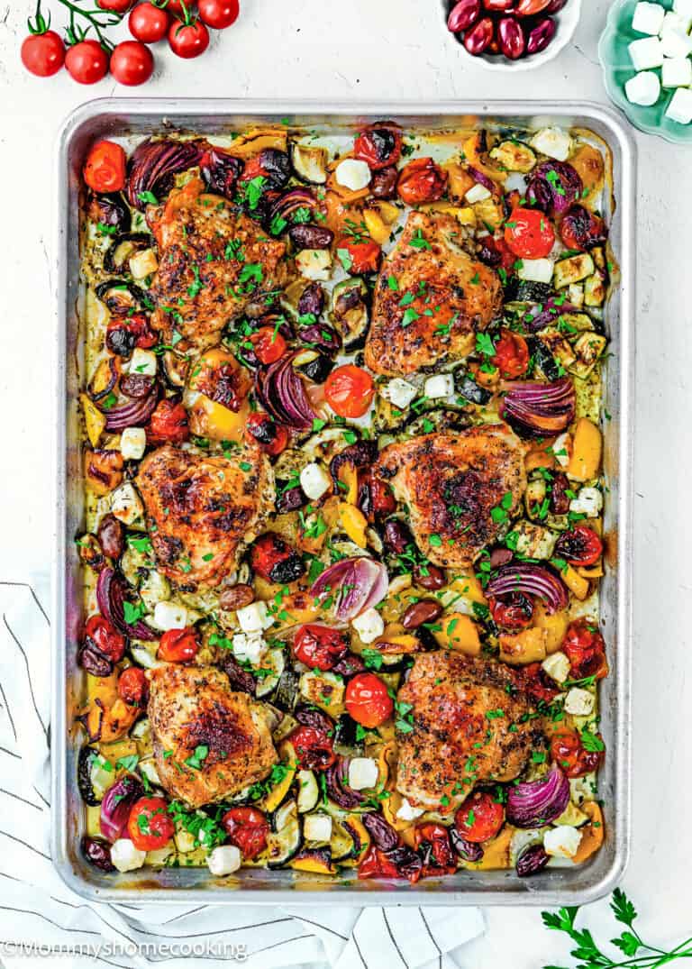 Easy Mediterranean Sheet-Pan Chicken: Roasted chicken thighs, red onions, zucchini, bell peppers, cherry tomatoes, and cubes of feta cheese garnished with fresh herbs—all on one pan for a delicious and effortless meal.