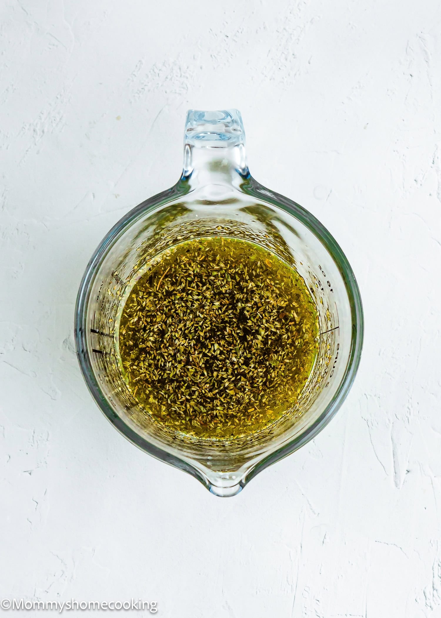 A glass measuring cup filled with a yellow-green liquid and visible dried herbs, perfect for marinating Easy Mediterranean Sheet-Pan Chicken, sits on a white textured surface.