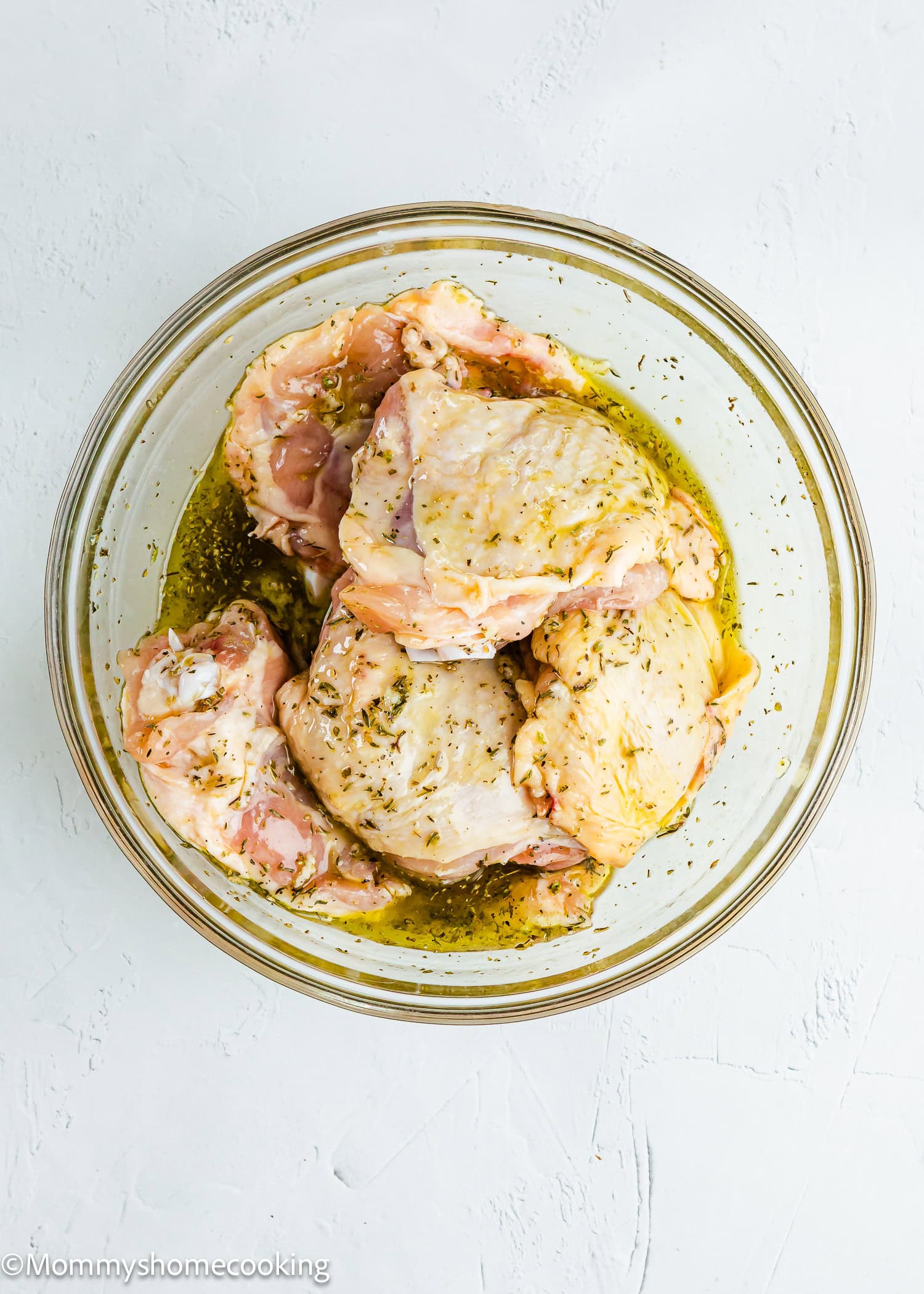Raw chicken pieces marinating in a clear glass bowl with oil and herbs on a white surface—perfect for making Easy Mediterranean Sheet-Pan Chicken.