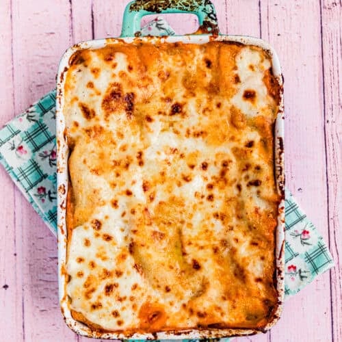 An easy Venezuelan Pasticho baked in a rectangular dish sits on a pink wooden surface, with a green patterned cloth underneath and fresh basil and cherry tomatoes nearby.