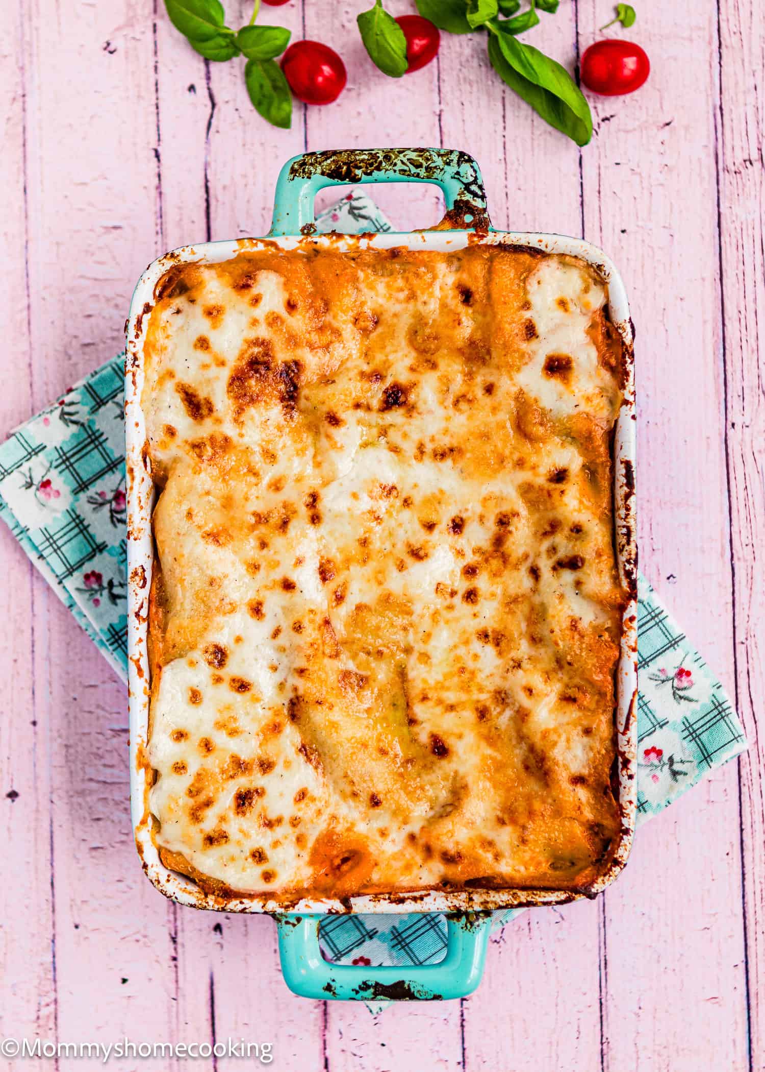 An easy Venezuelan Pasticho baked in a rectangular dish sits on a pink wooden surface, with a green patterned cloth underneath and fresh basil and cherry tomatoes nearby.