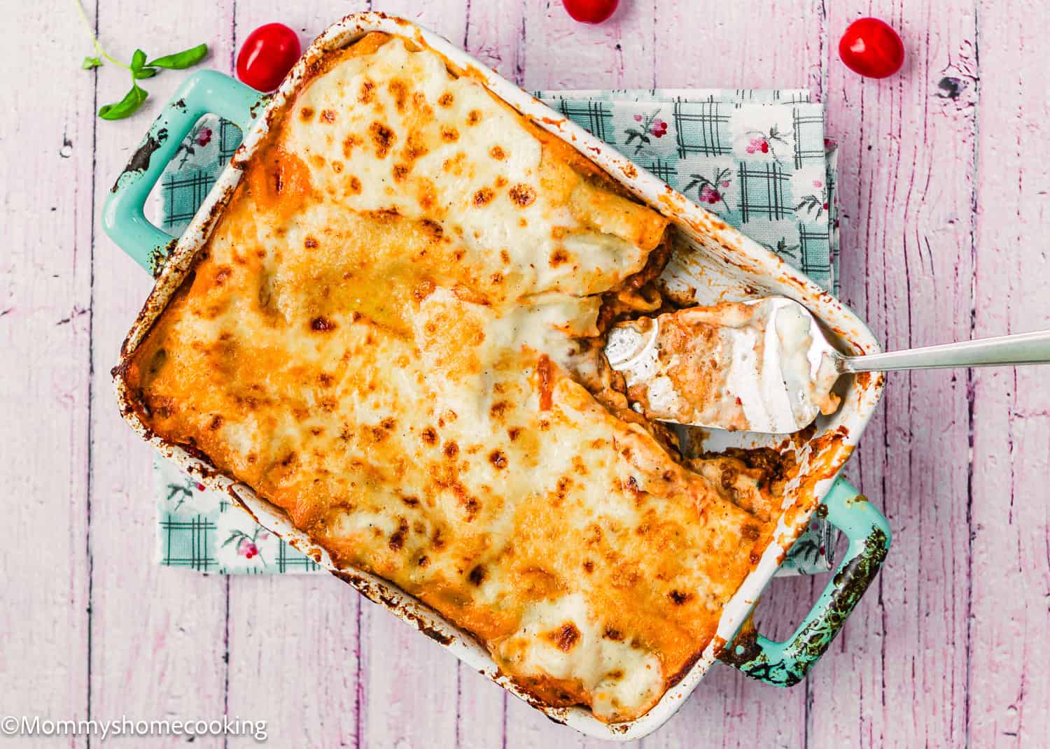 Easy Venezuelan Pasticho baked in a casserole dish with a serving removed; spatula rests inside. The dish sits on a patterned cloth next to cherry tomatoes on a pink wooden surface.