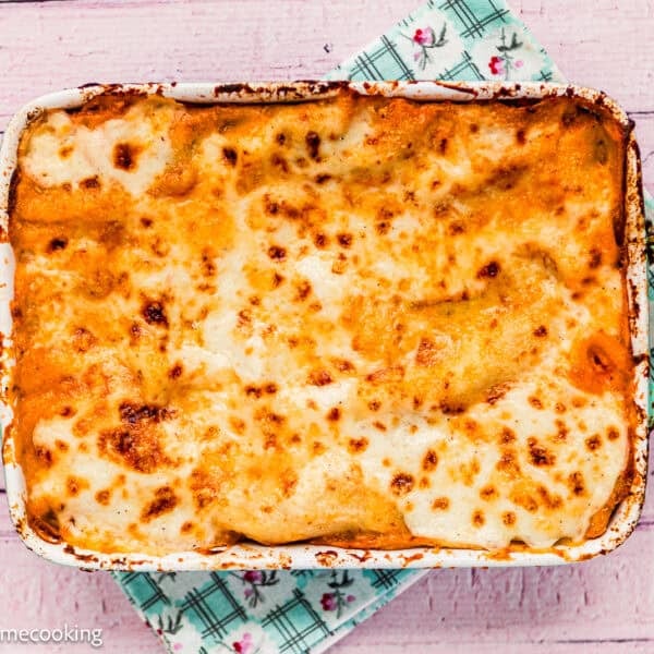 Baked lasagna in a rectangular green dish, topped with melted and slightly browned cheese, rests on a floral-patterned cloth on a pink wooden surface—reminiscent of an easy Venezuelan Pasticho.