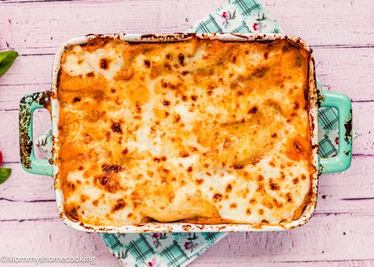 Baked lasagna in a rectangular green dish, topped with melted and slightly browned cheese, rests on a floral-patterned cloth on a pink wooden surface—reminiscent of an easy Venezuelan Pasticho.