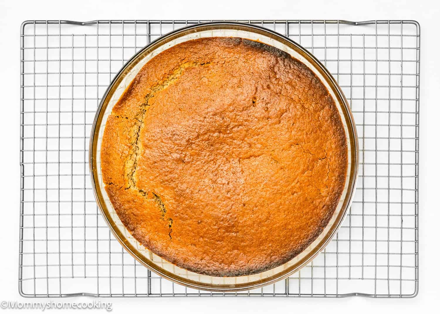 A round, golden-brown Eggless Easy Apple Upside Down Cake in a glass dish sits on a cooling rack against a white background.