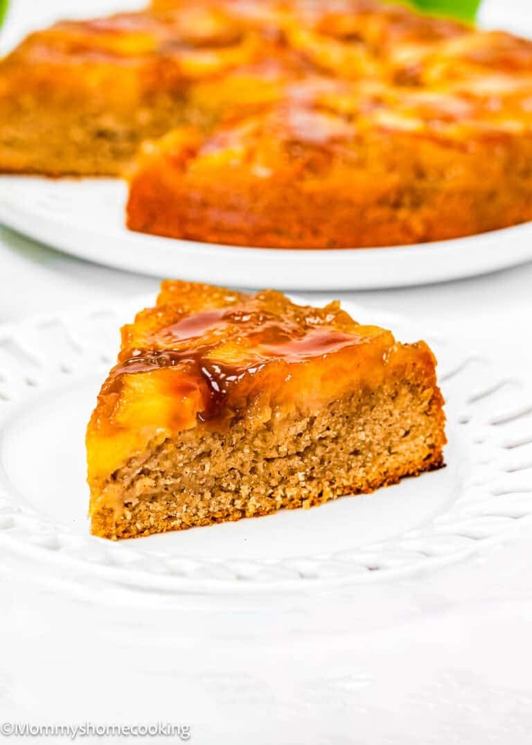 A slice of pineapple upside-down cake on a white plate with the rest of the Eggless Easy Apple Upside Down Cake in the background.