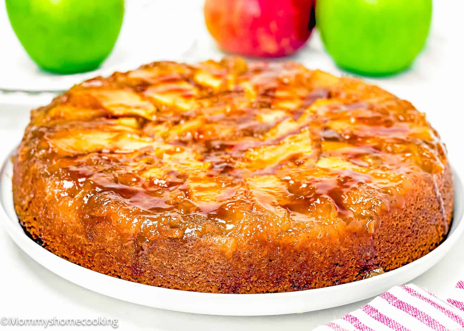A round Eggless Easy Apple Upside Down Cake sits on a white plate, its glazed apple slices gleaming on top. Two apples are blurred in the background.
