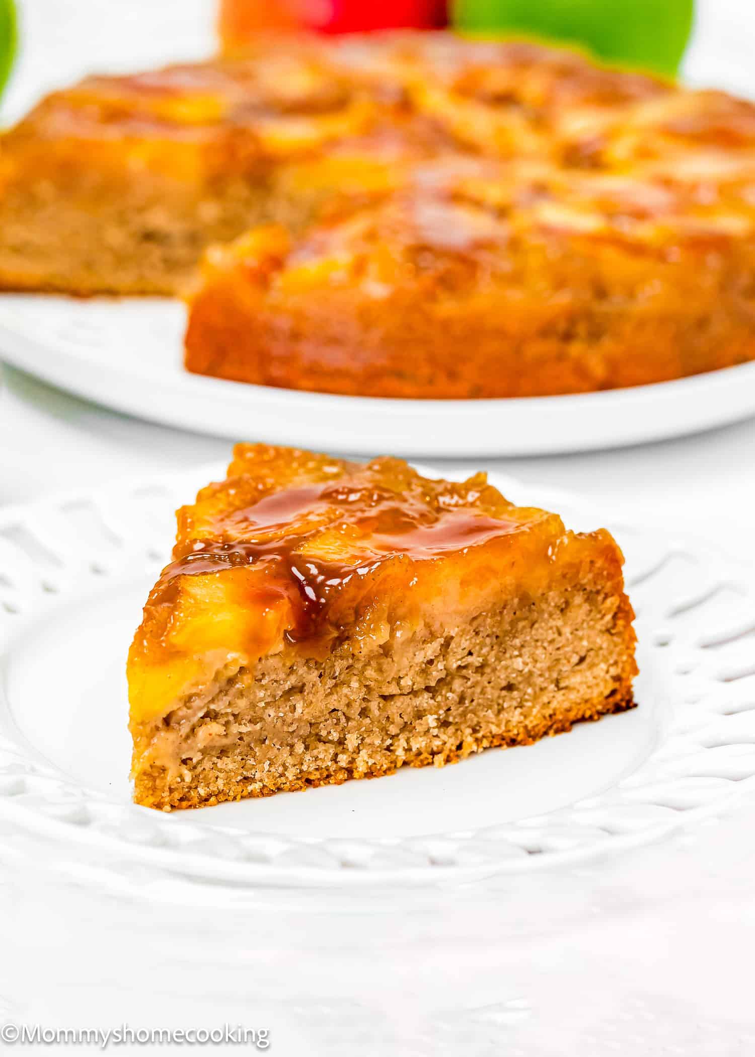 A slice of pineapple upside-down cake on a white plate with the rest of the Eggless Easy Apple Upside Down Cake in the background.