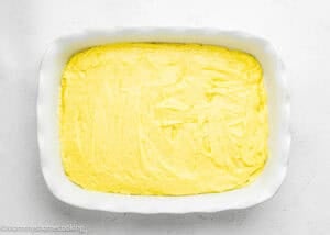 Rectangular white baking dish filled with an even layer of uncooked Eggless Lemon Poke Cake batter on a white surface.