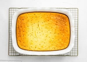 An Eggless Lemon Poke Cake, rectangular and baked to perfection, features small holes on its surface and sits in a white dish on a cooling rack.
