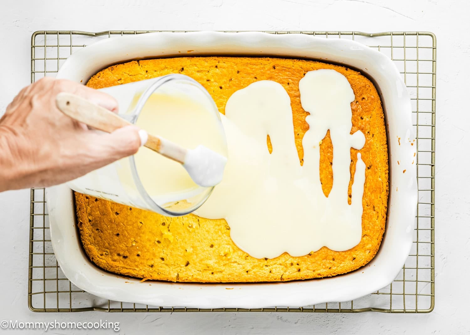 A hand pours white icing over an Eggless Lemon Poke Cake in a rectangular white baking dish on a wire rack.
