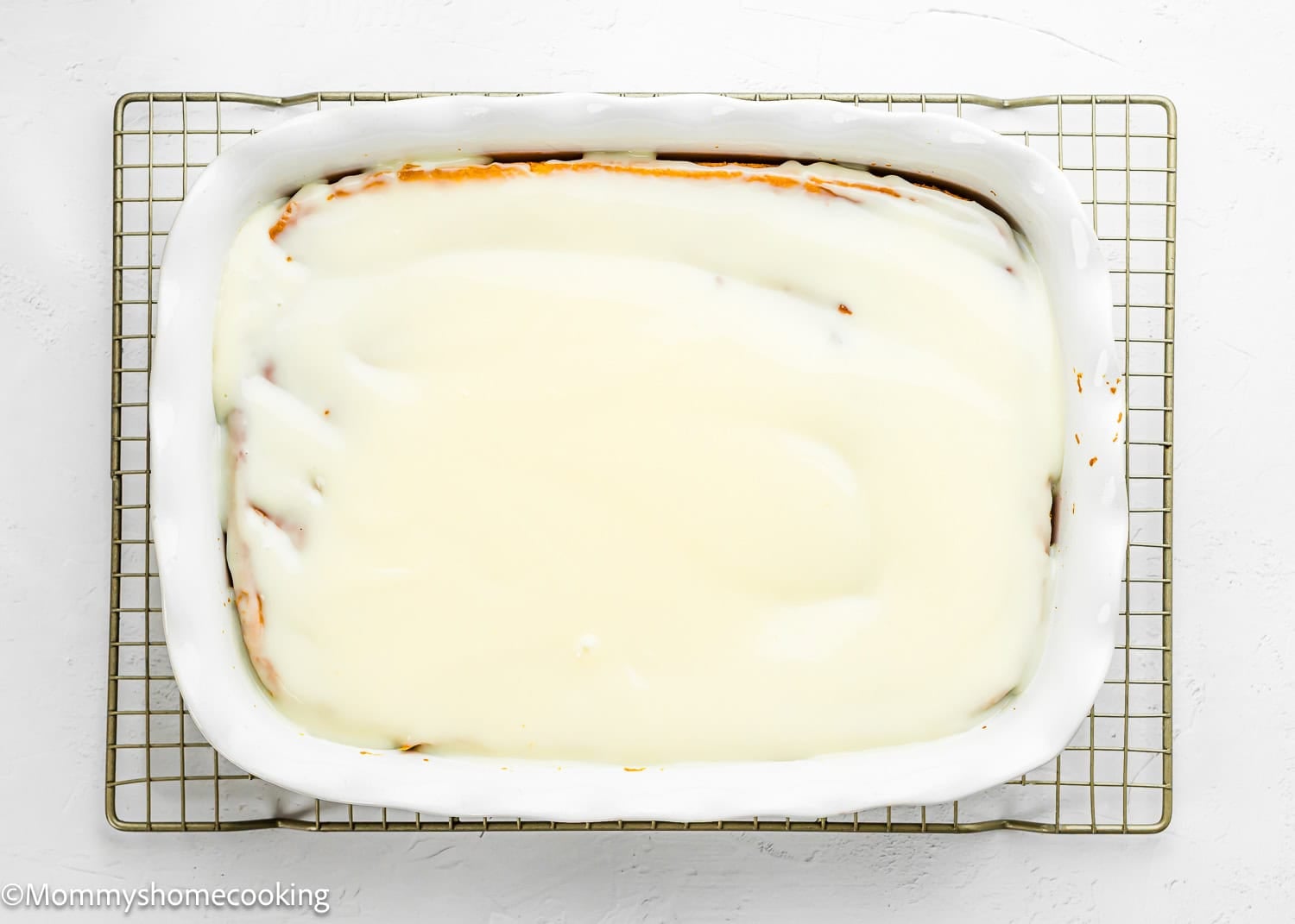 A rectangular Eggless Lemon Poke Cake in a white baking dish is covered with a smooth layer of lemon sauce, resting on a wire cooling rack.