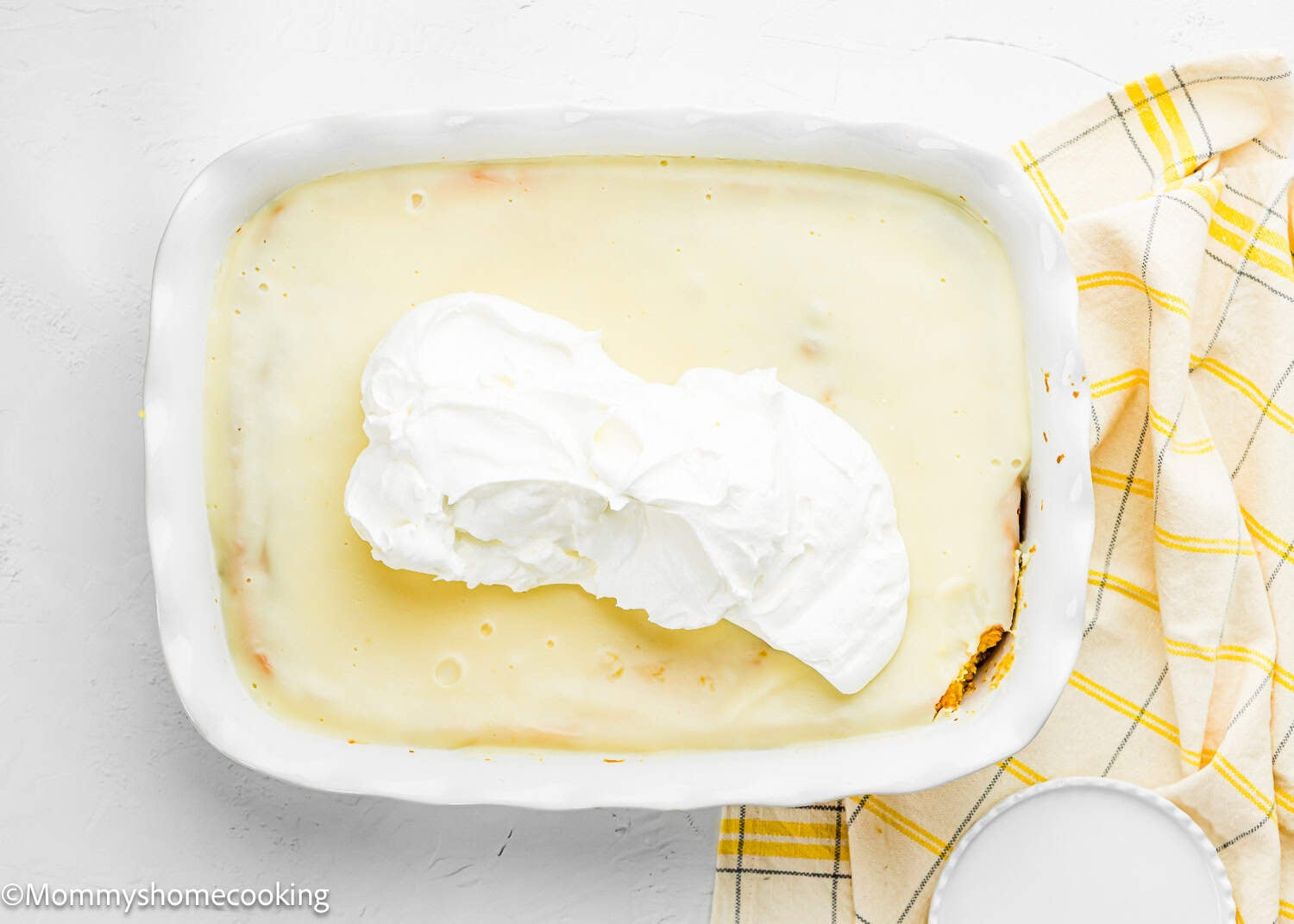 A rectangular baking dish holds a creamy Eggless Lemon Poke Cake, crowned with a generous dollop of whipped cream, set beside a yellow plaid kitchen towel.