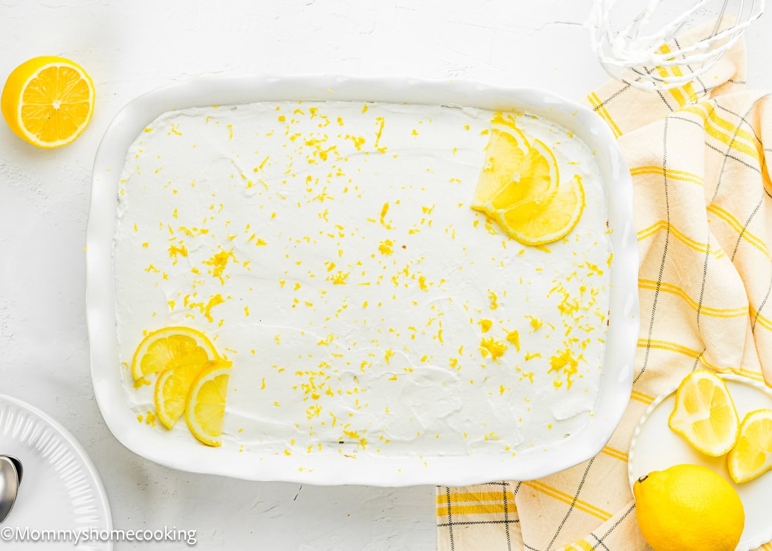 Rectangular dish filled with a white, frosted Eggless Lemon Poke Cake topped with lemon zest and slices, placed on a white surface with a yellow checked cloth, lemon, and plated dessert nearby.