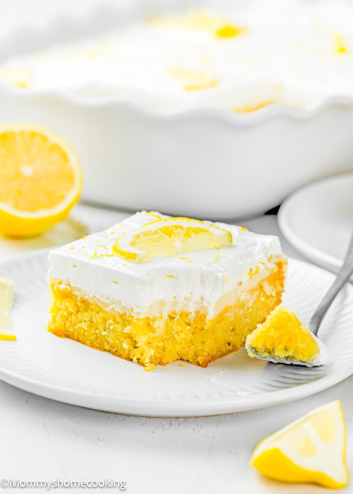 A slice of Eggless Lemon Poke Cake with white frosting, topped with a lemon slice, sits on a white plate with a fork and lemon wedges nearby.