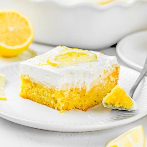A slice of Eggless Lemon Poke Cake with white frosting, topped with a lemon slice, sits on a white plate with a fork and lemon wedges nearby.