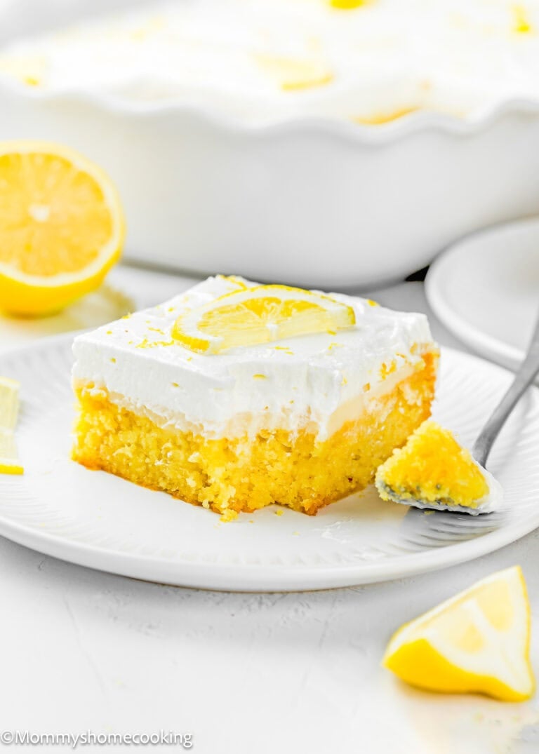 A slice of Eggless Lemon Poke Cake with white frosting, topped with a lemon slice, sits on a white plate with a fork and lemon wedges nearby.