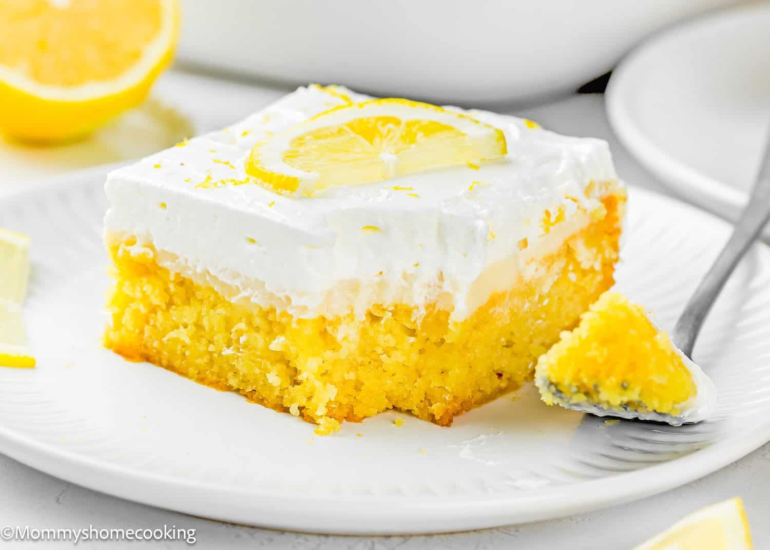 A slice of Eggless Lemon Poke Cake with creamy white frosting and a lemon wedge on top sits on a white plate, with a fork holding a bite-sized piece next to it.