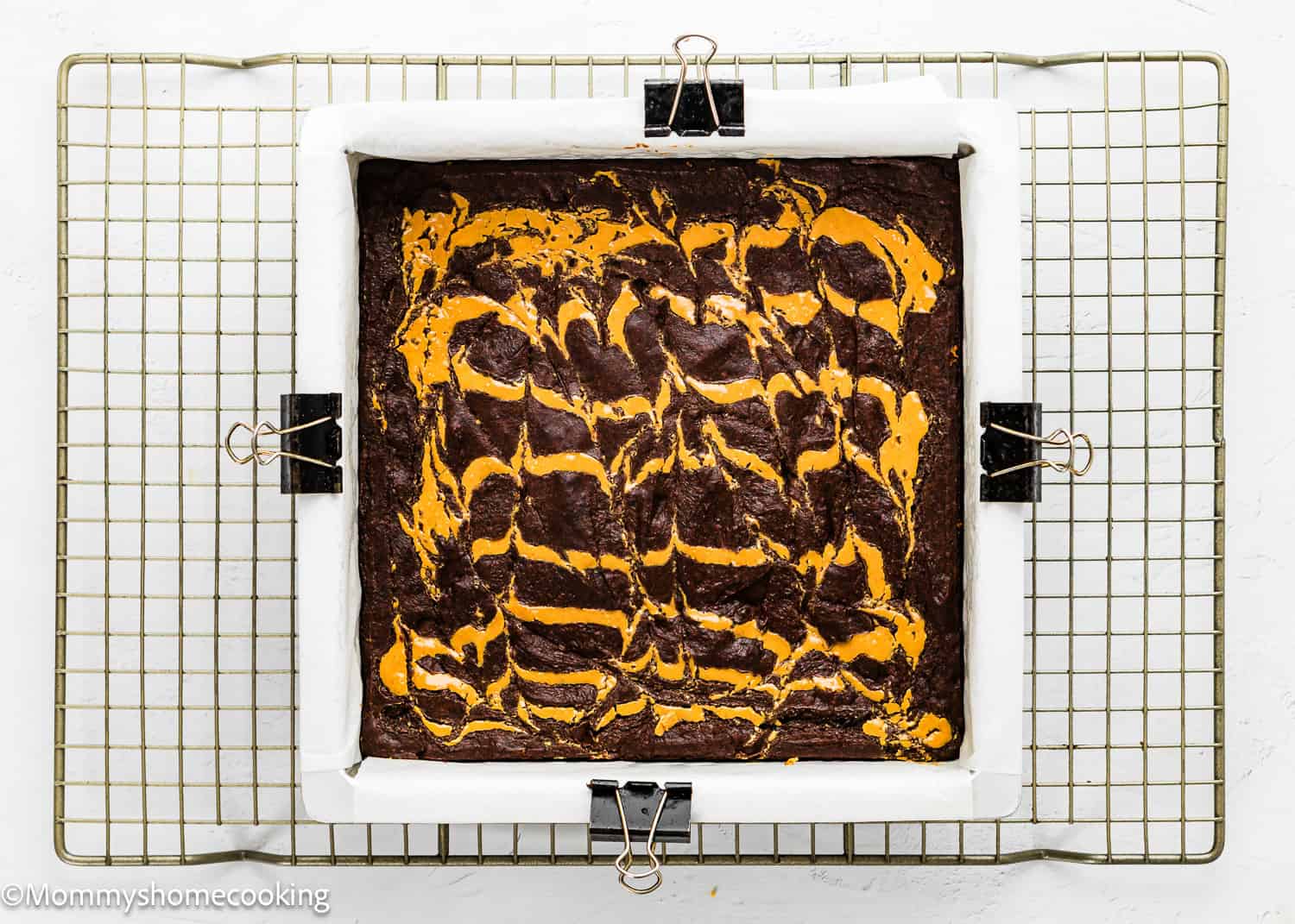 A square pan of chocolate brownies with orange swirled topping sits on a cooling rack, lined with parchment paper and secured with binder clips.