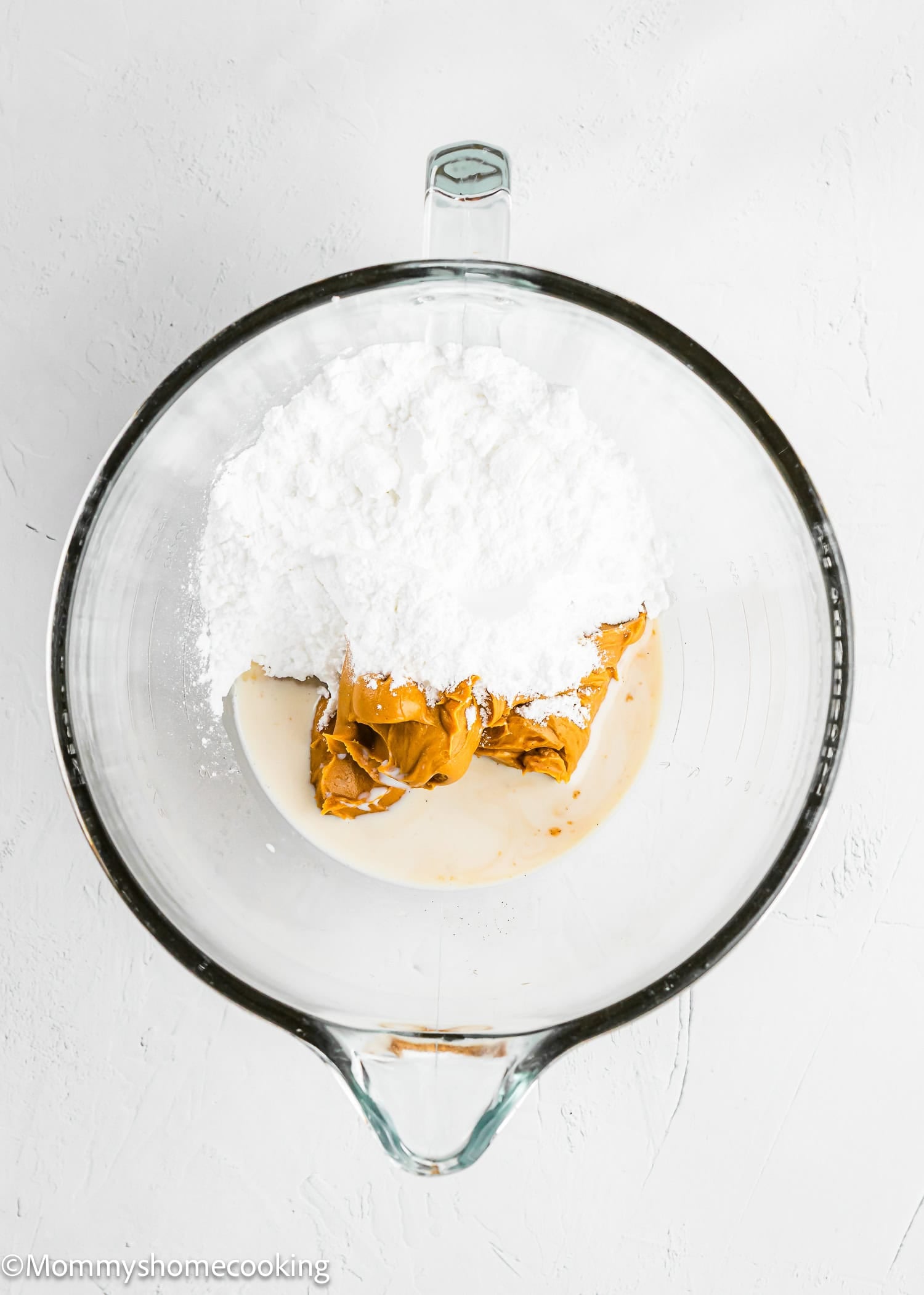 A glass mixing bowl containing powdered sugar, peanut butter, and a small amount of liquid on a white surface.