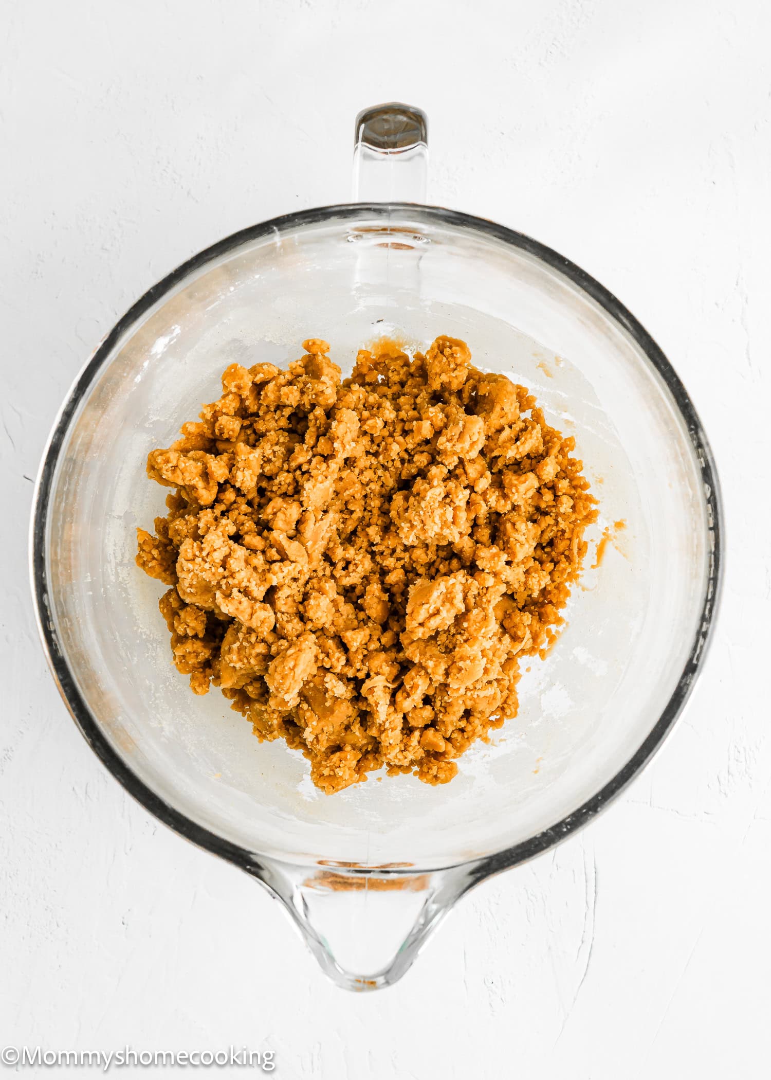 A glass mixing bowl containing a clump of light brown cookie dough for eggless peanut butter brownies rests on a white surface.
