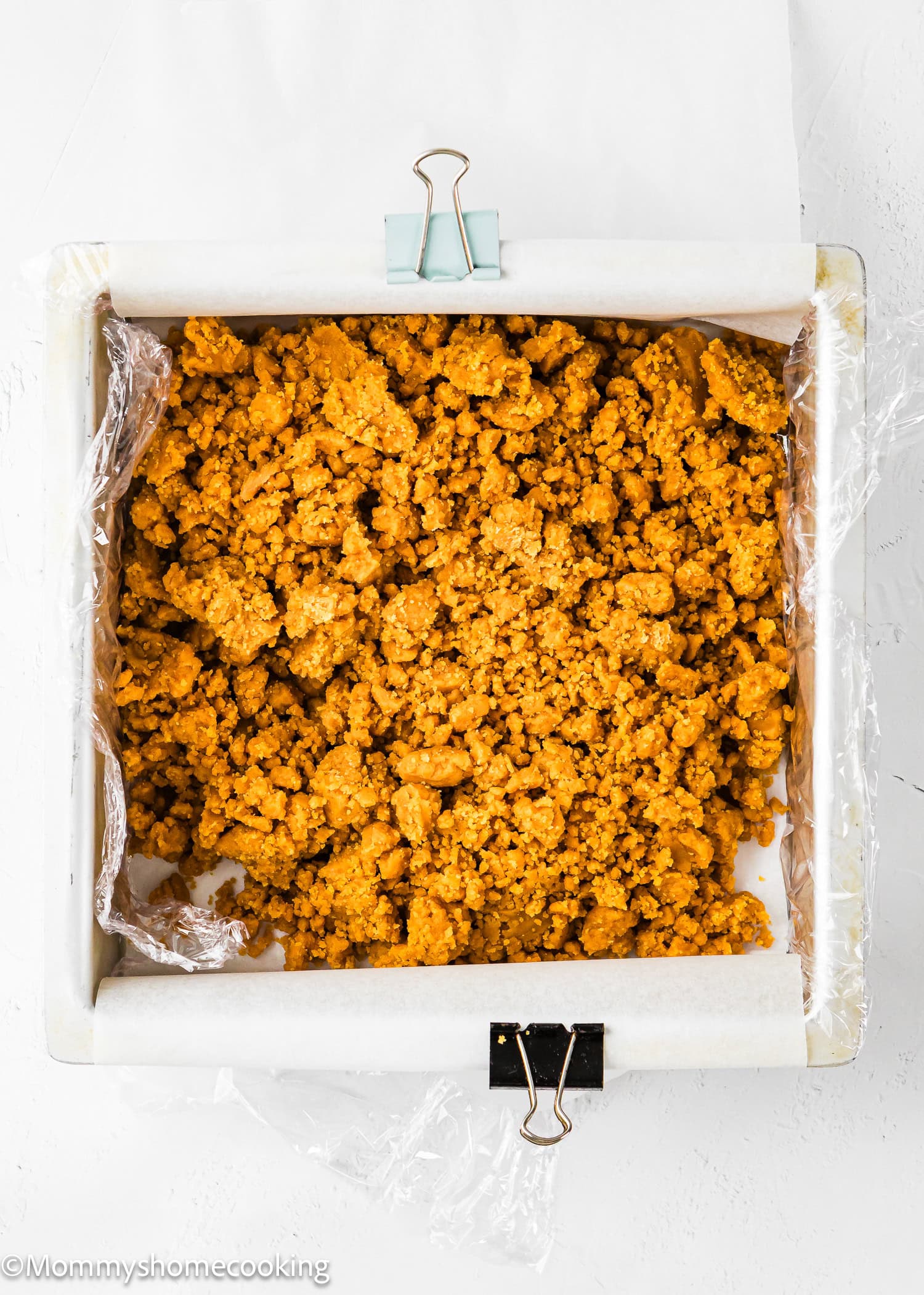 A baking pan lined with parchment paper and plastic wrap, filled with a crumbly orange mixture, secured with binder clips on two sides.