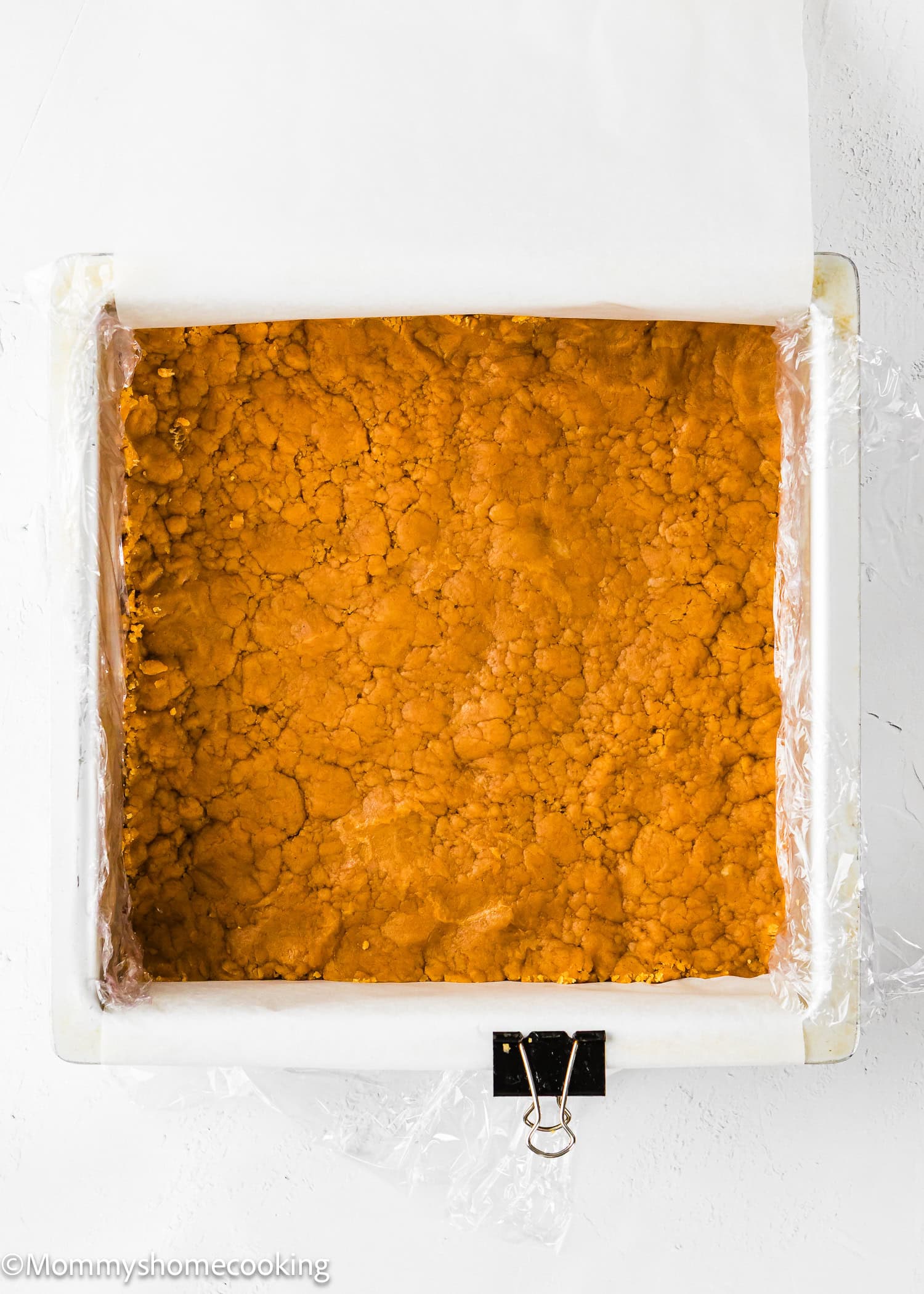 A square baking pan lined with parchment paper and filled with a pressed, crumbly orange mixture, secured with a binder clip on one side.