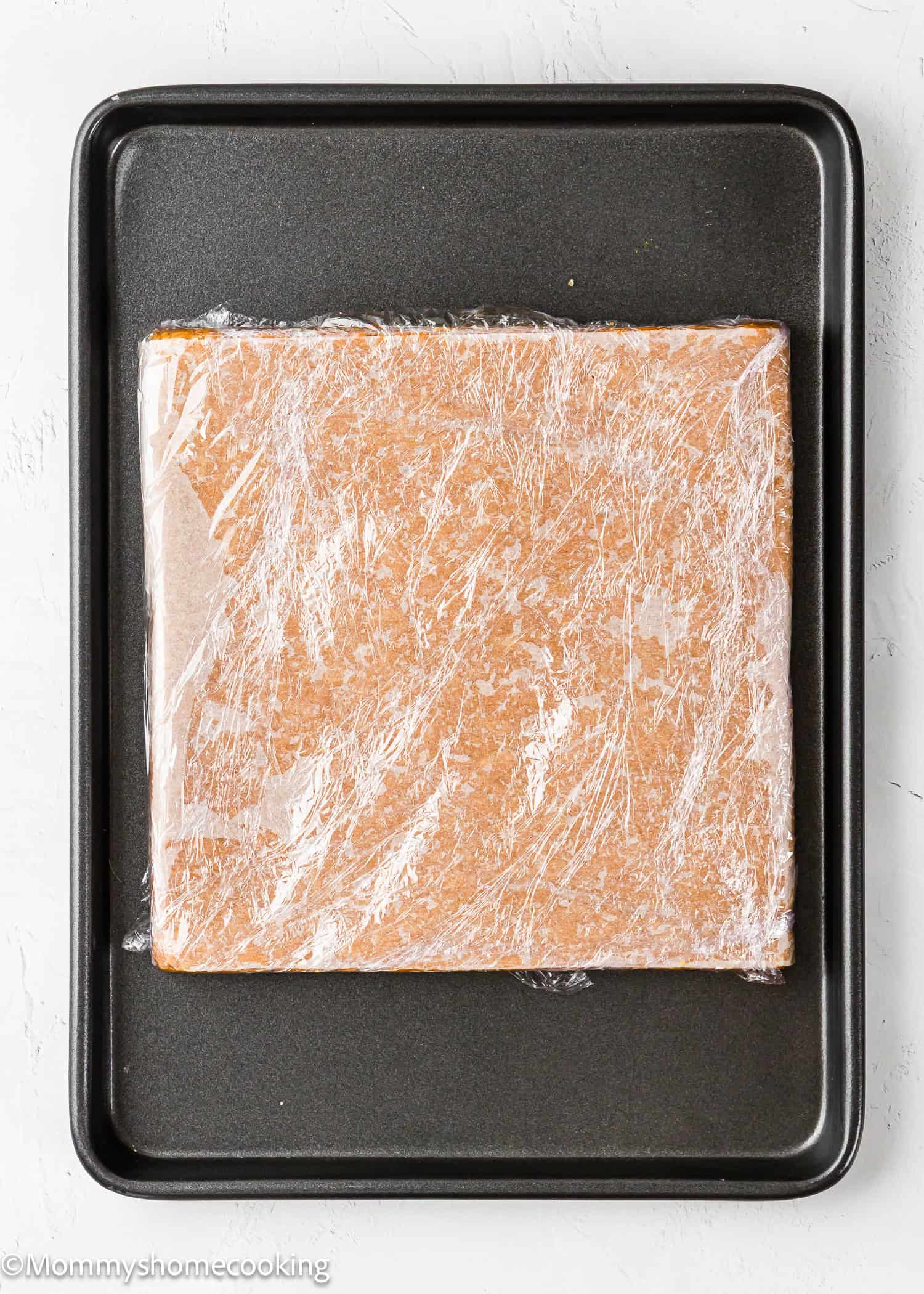 A square block of dough wrapped in plastic wrap sits on a black baking tray.