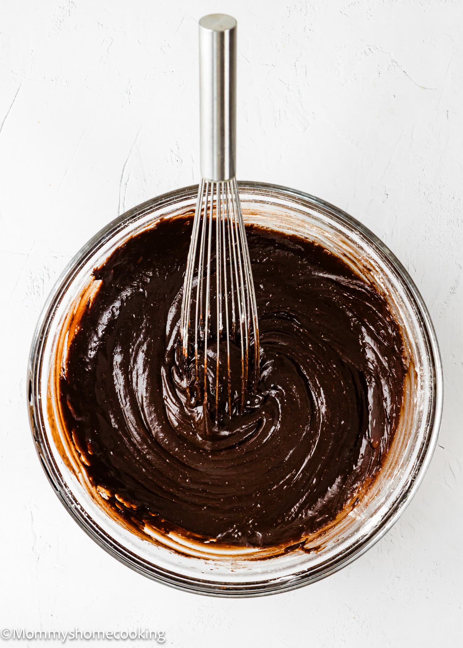 A metal whisk stirs thick chocolate brownie batter in a clear glass bowl on a white surface.