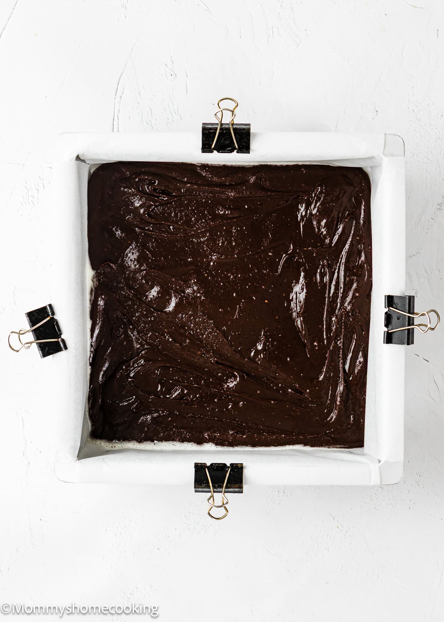 Square baking pan lined with parchment paper and binder clips, filled with unbaked chocolate batter, on a white surface.