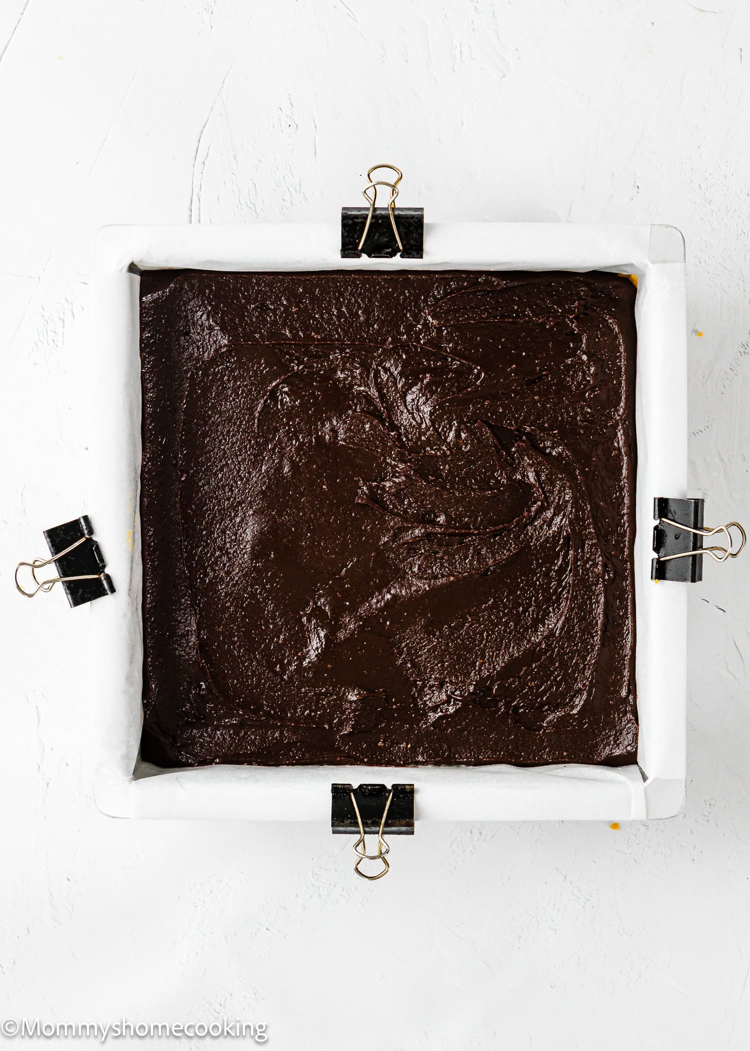 A square baking pan lined with parchment paper and binder clips, filled with unbaked chocolate batter, sits on a white surface.